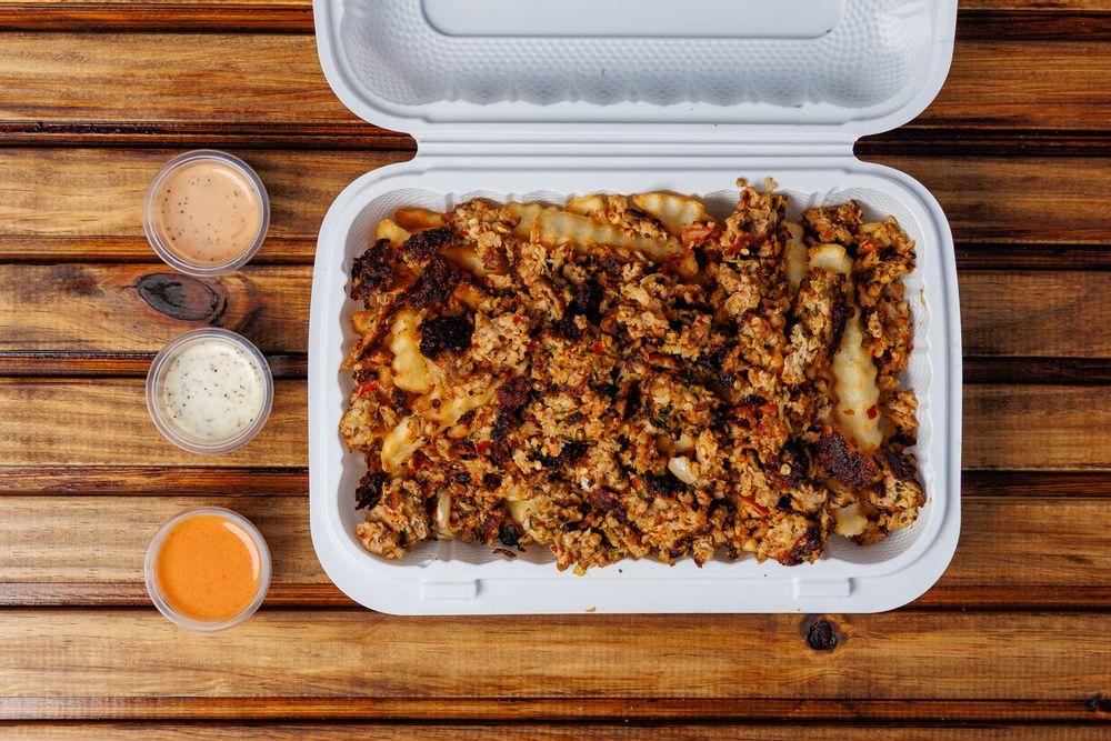A takeaway box of crinkle-cut fries covered in meat, with three sauce cups alongside.