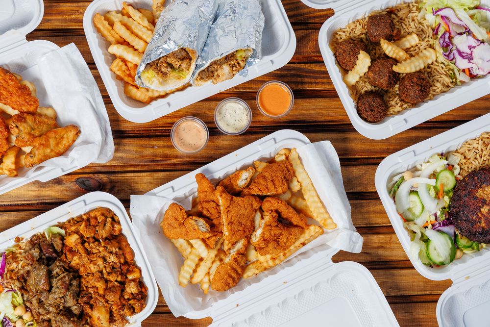 An overhead spread of Khan Baba takeaway boxes: wings, a wrap with fries, chicken tenders, rice platters and three sauce cups on a wooden table.