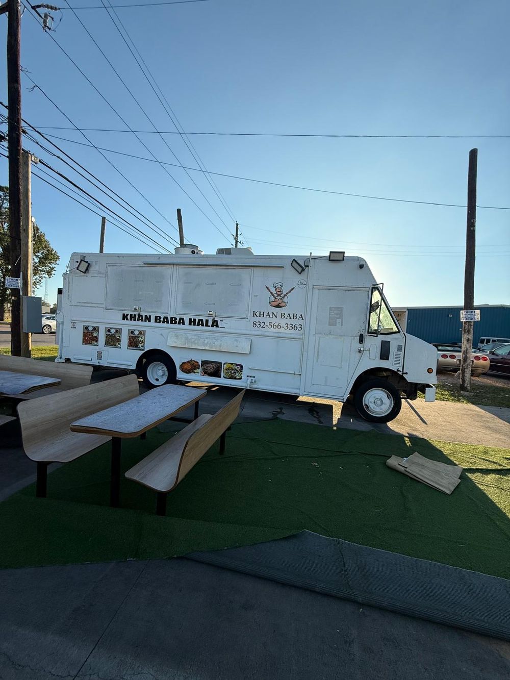 Picnic tables and benches on artificial grass beside the Khan Baba food truck in daylight.